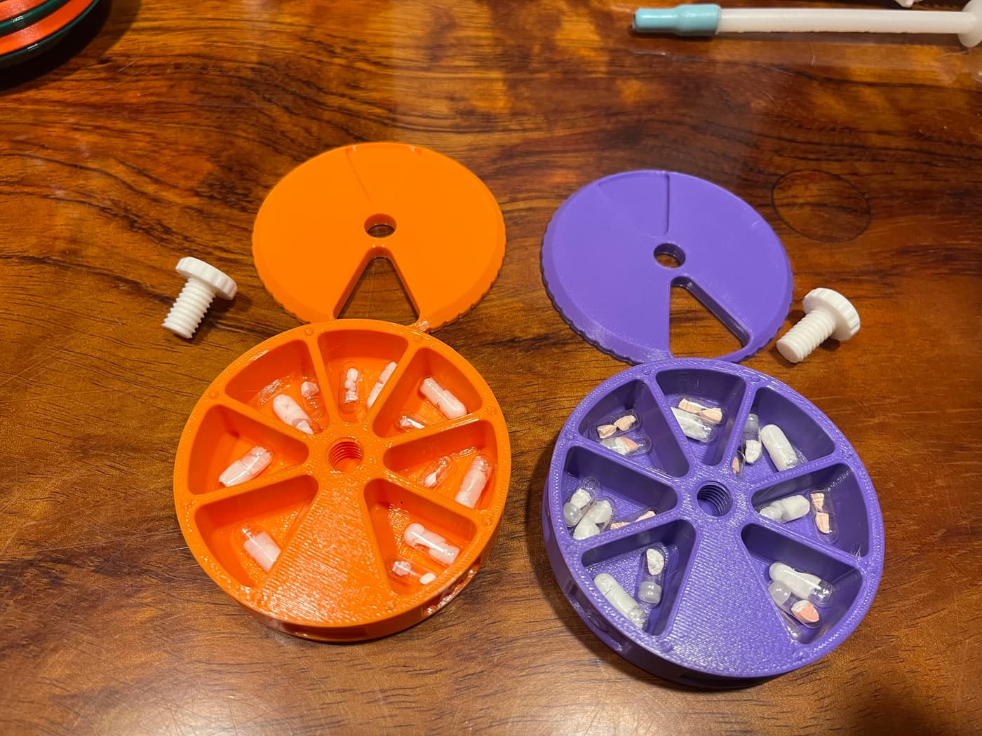 Two circular containers, one orange and one purple, are displayed on a wooden surface. Each container has compartments filled with small variously colored pills. A white plastic screw sits beside each container.