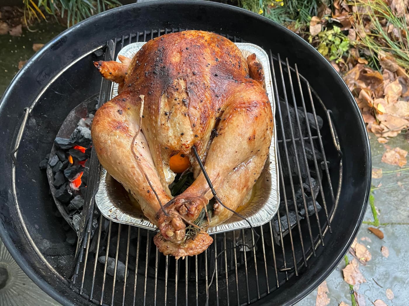 A roasted turkey is placed in a grill over charcoal. The turkey has a golden-brown skin and is resting in a foil tray, with some herbs visible inside. Surrounding the grill are fallen leaves and greenery.