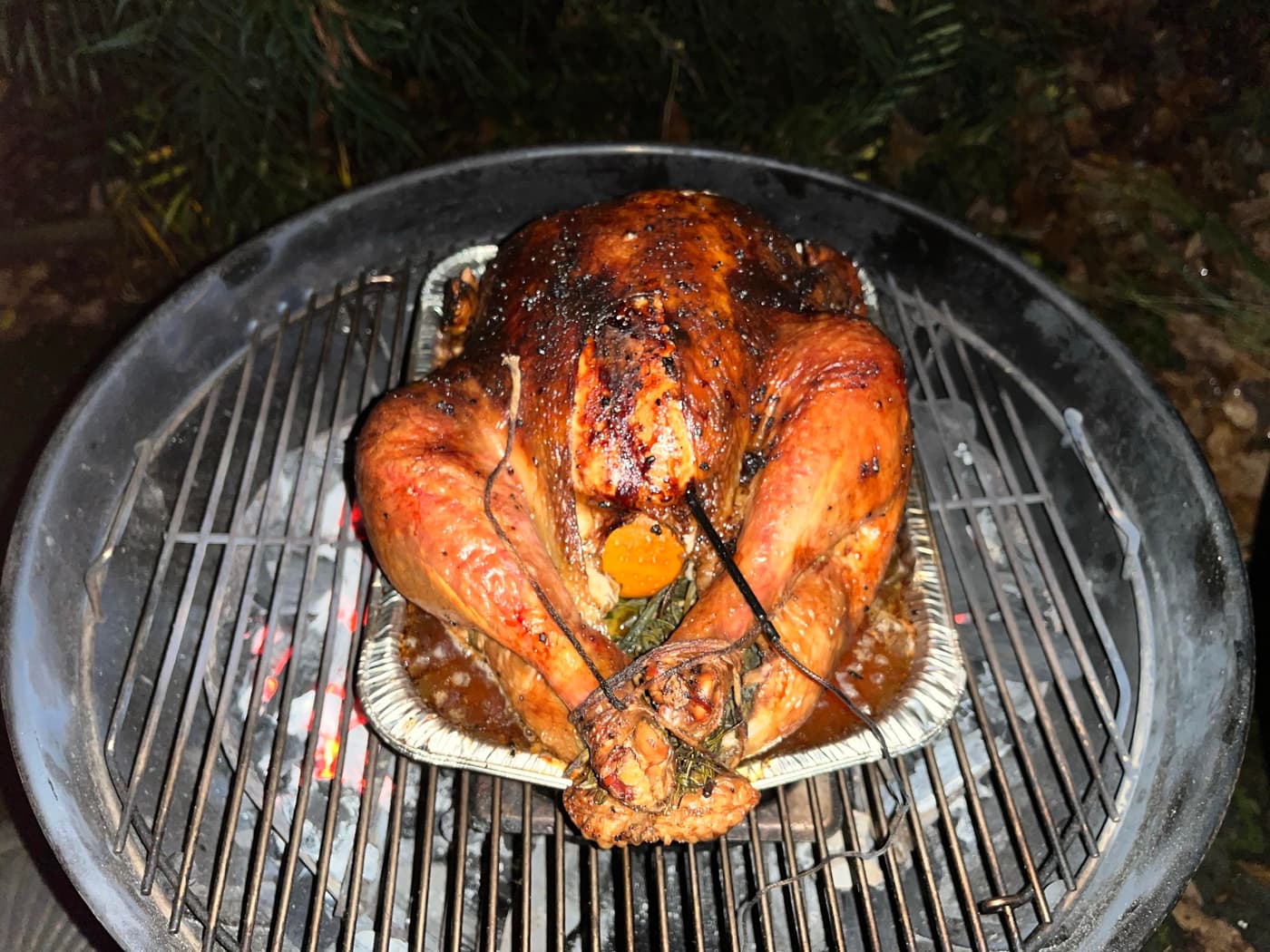 A whole turkey is being grilled over charcoal, resting in a metal tray. The turkey has a shiny, glazed skin and is surrounded by smoke, with a dark, outdoor setting in the background.