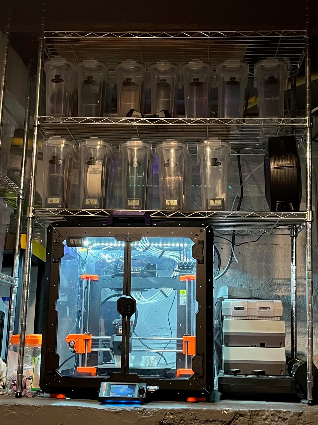 Photo of a prusa MK4S in an enclosure, under a steel shelving unit with multiple plastic canisters containing spools of 3D printer filament