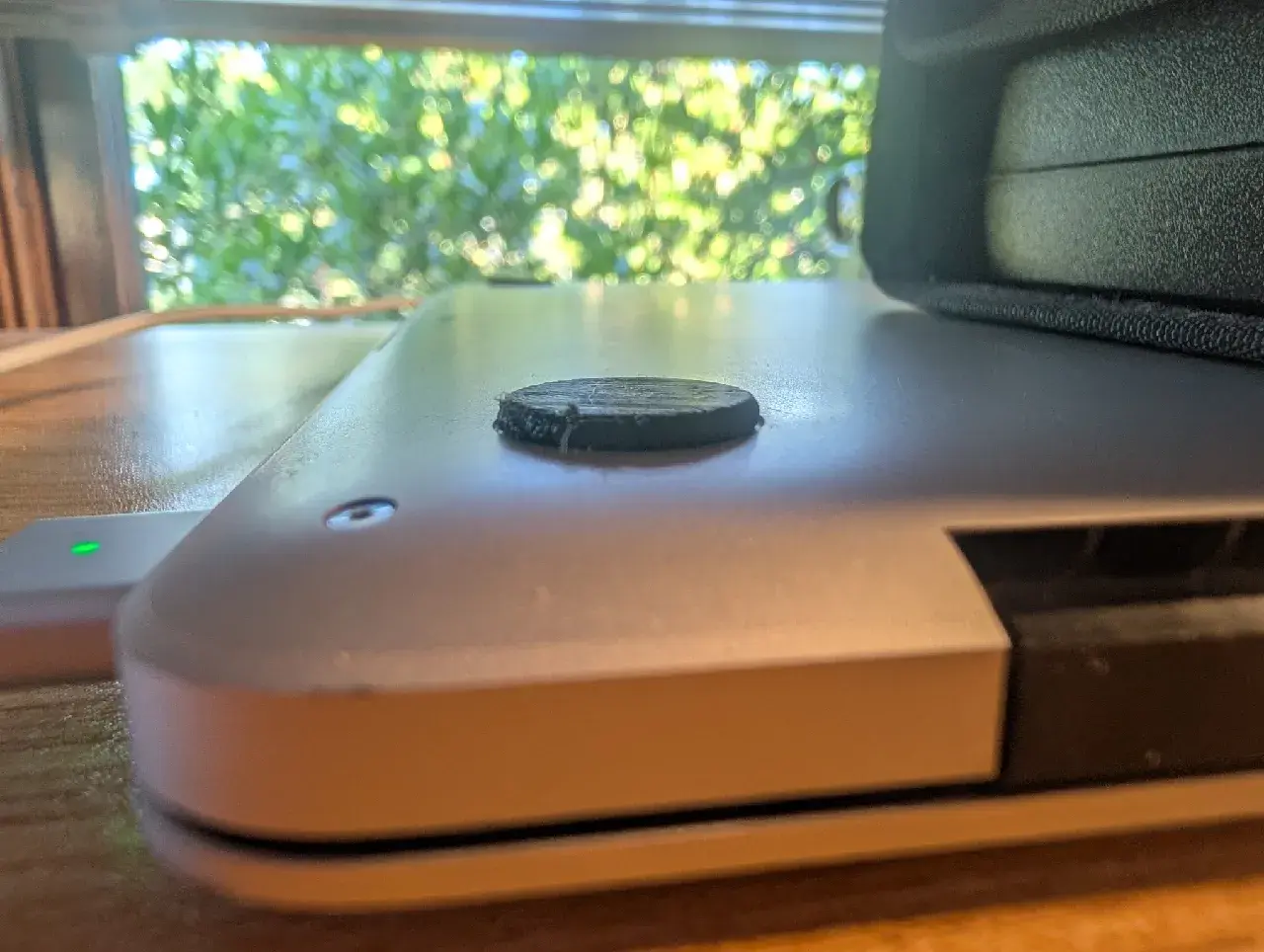 Close-up of one corner of the MacBook's underside, with a black 3D-printed disc foot stuck on and green trees blurred through a window behind