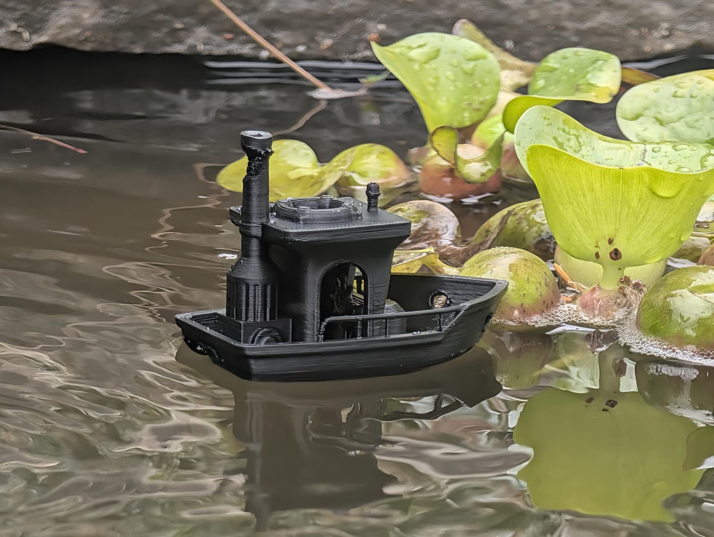 A black 3D-printed tugboat floating on a pond next to green water hyacinths