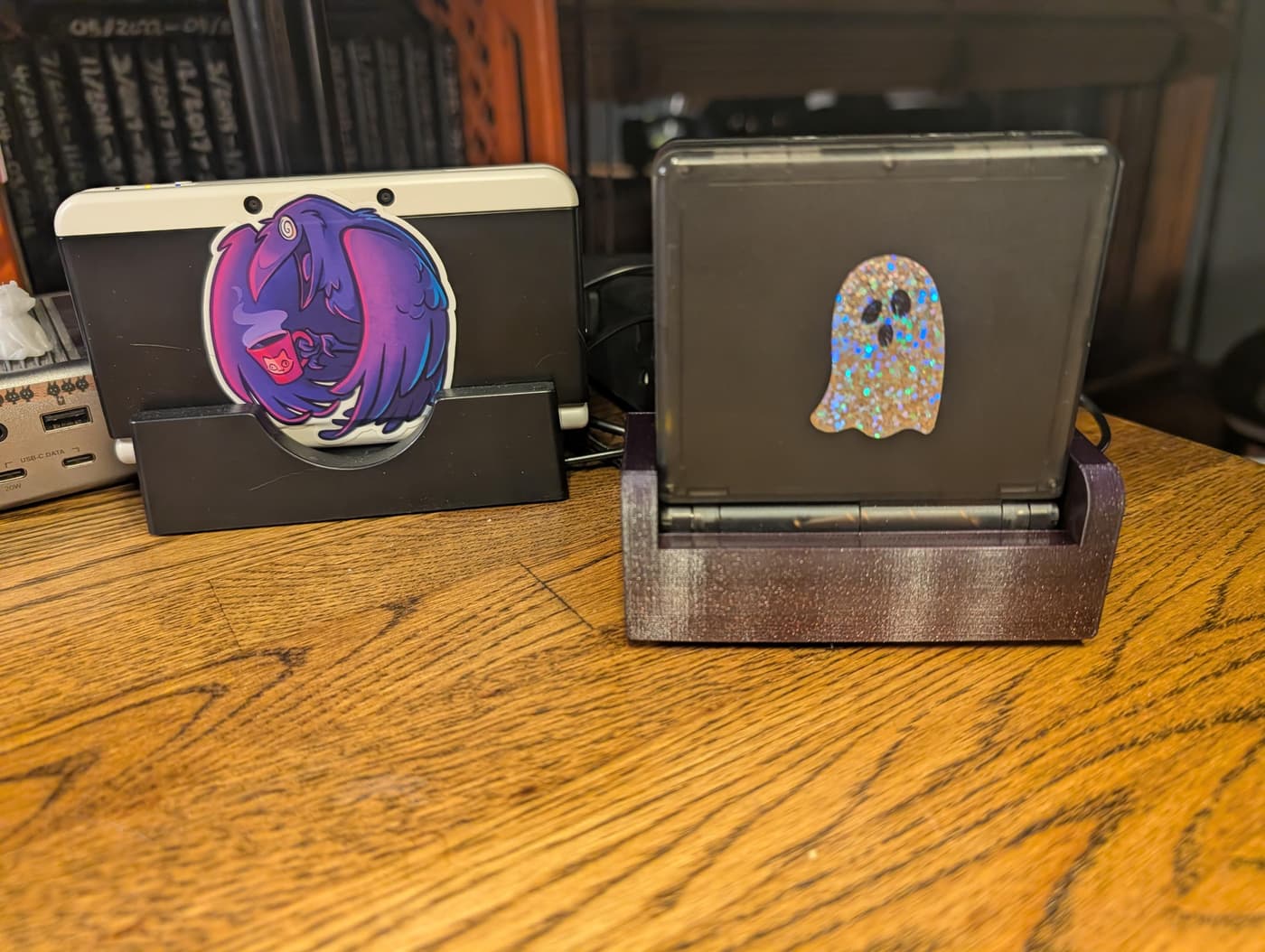 Two handheld consoles, a New Nintendo 3DS with a purple bird sticker and an Anbernic 35xxsp with a glittery ghost sticker, sit in stands on a wooden desk.